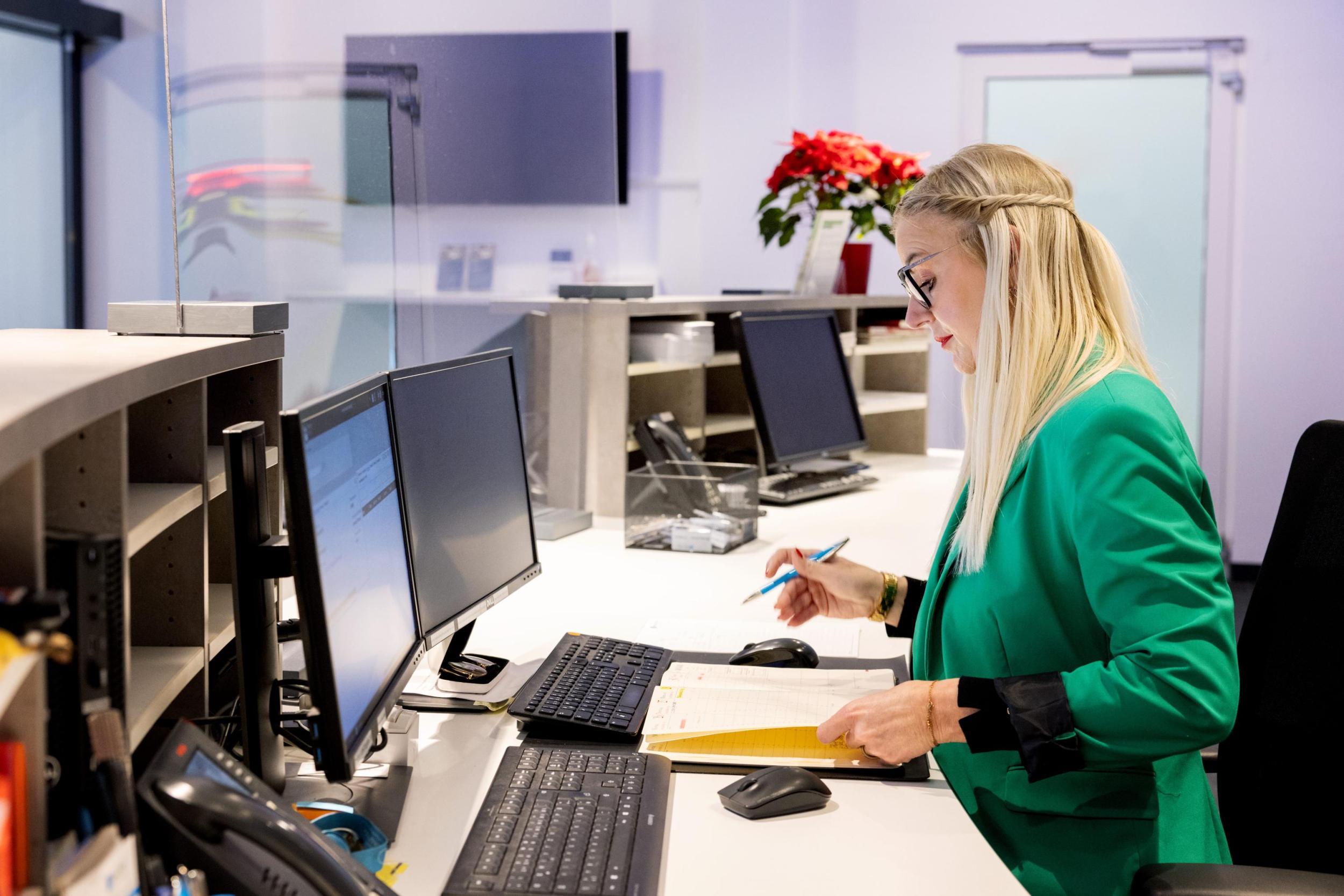 women wearing a green jacket, sitting behind the counter in front of computer, writing on note pad