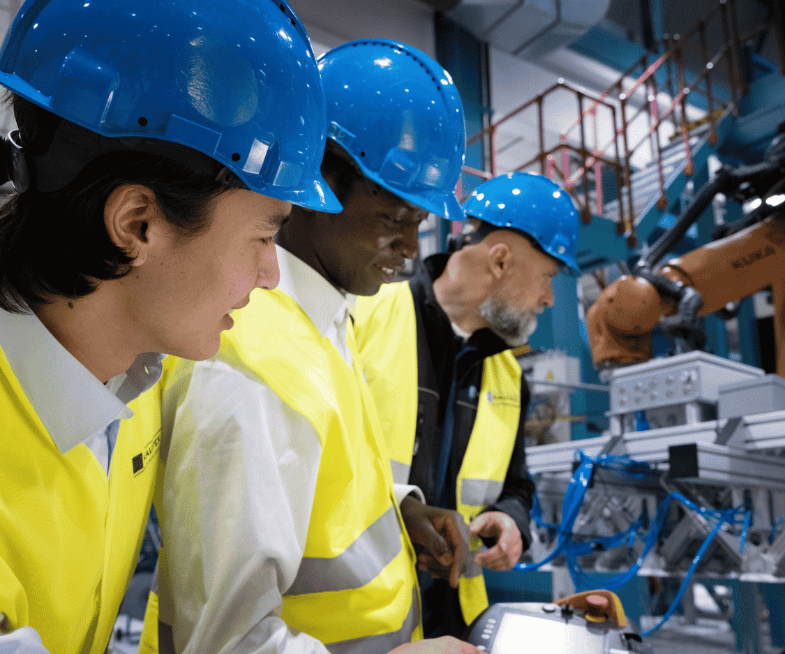Three employees wearing blue hard hats and high-visibility safety vests work alongside automated equipment in a manufacturing facility.