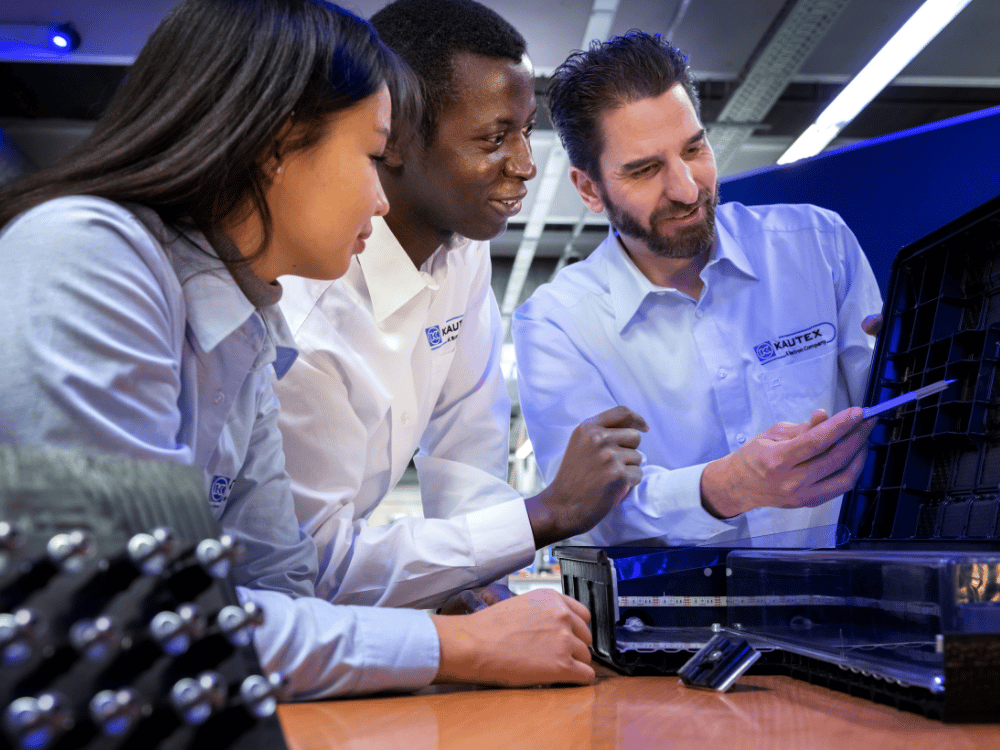 Three employees wearing Kautex-branded shirts are gathered around a workstation, examining a molded plastic component while one uses a pen to point out details on the part, with tools and equipment visible on the table in a modern lab or office setting.